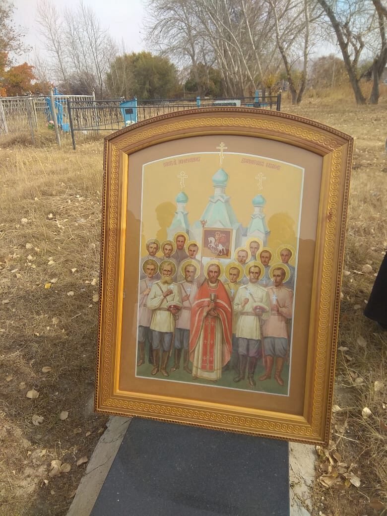 Икона собора новомучеников в киоте, стоит на плите памятника: священник в красном облачении с потиром, вокруг около четырнадцати мирян в русских рубахах, на фоне храм с бирюзовыми куполами